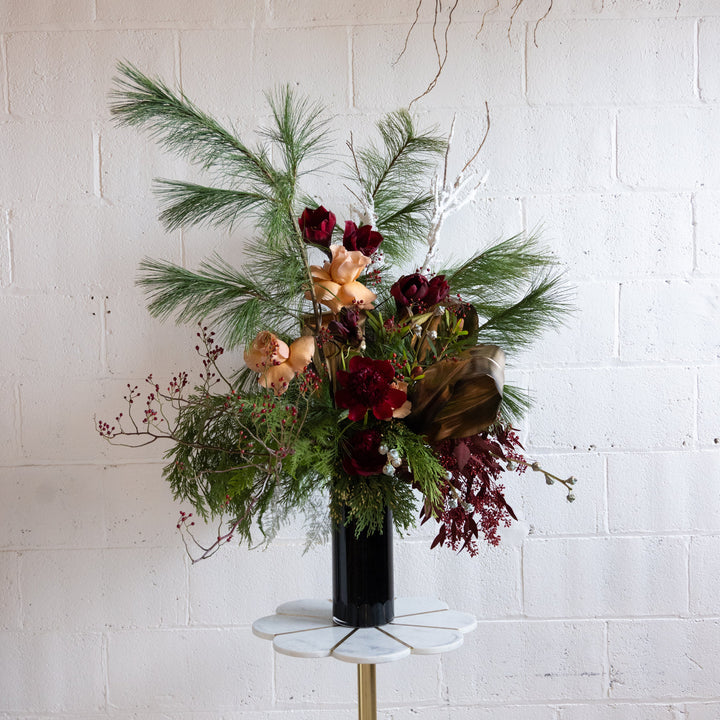 Floral arrangement with greenery and red flowers in a black vase on a white surface against a white brick wall.