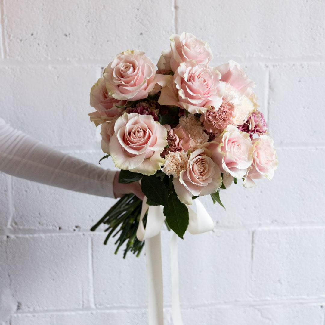 Bouquet of pink and white roses held by a person against a white brick wall.