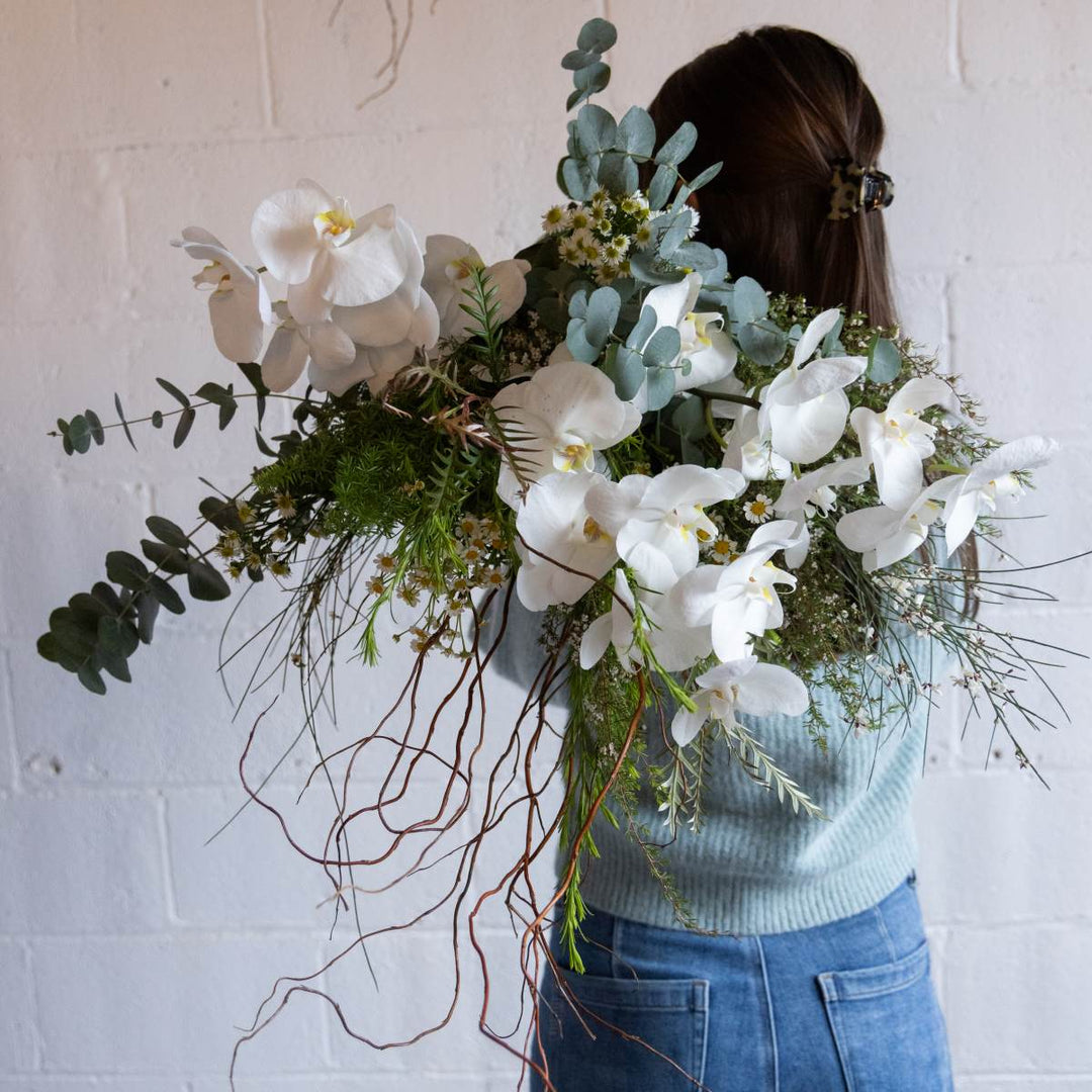 Person holding a large bouquet of white orchids and greenery against a white wall.
