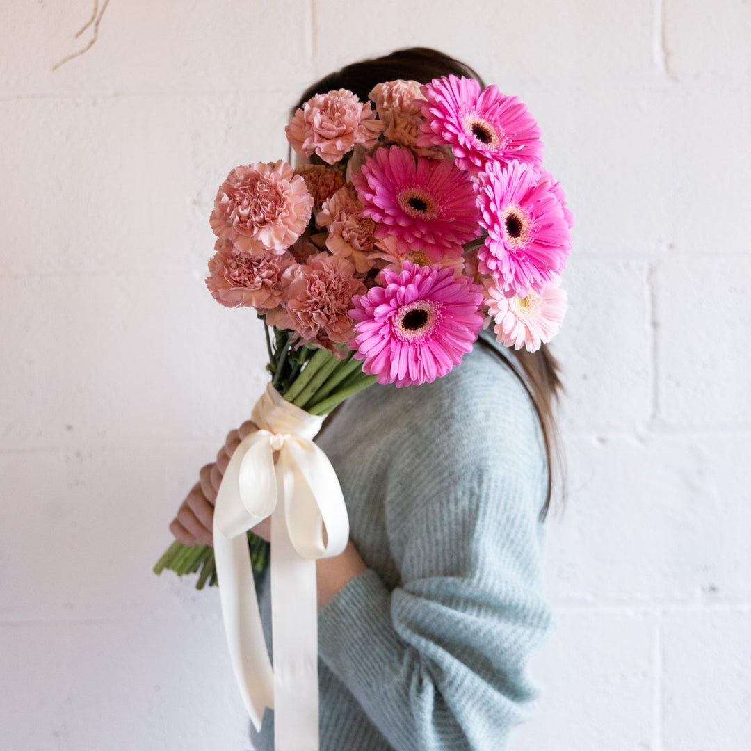 Person holding a bouquet of pink and peach flowers with a white ribbon against a light background