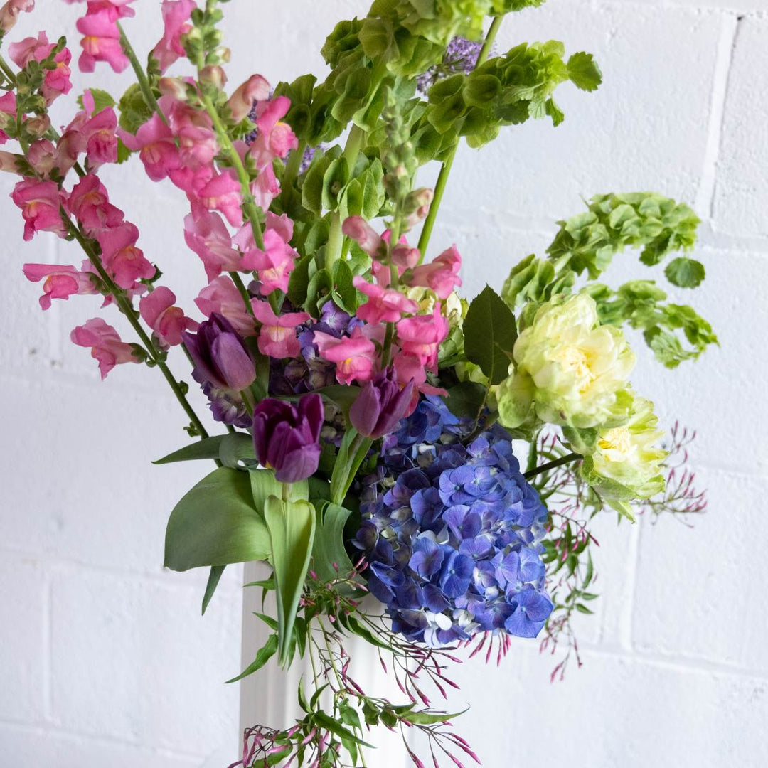 Bouquet of colorful flowers including pink, purple, and blue against a white brick wall.