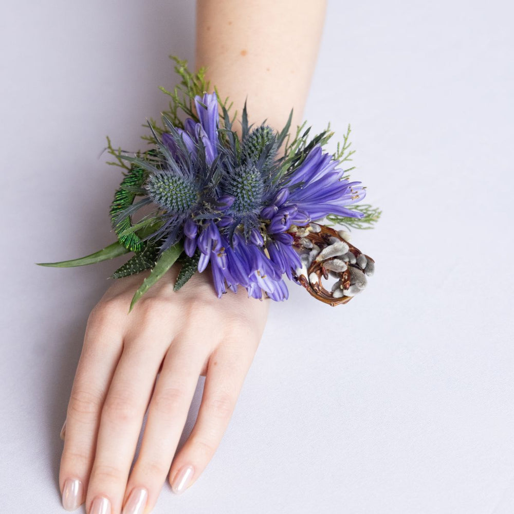 Hand wearing a wrist corsage with purple flowers and greenery on a light gray background