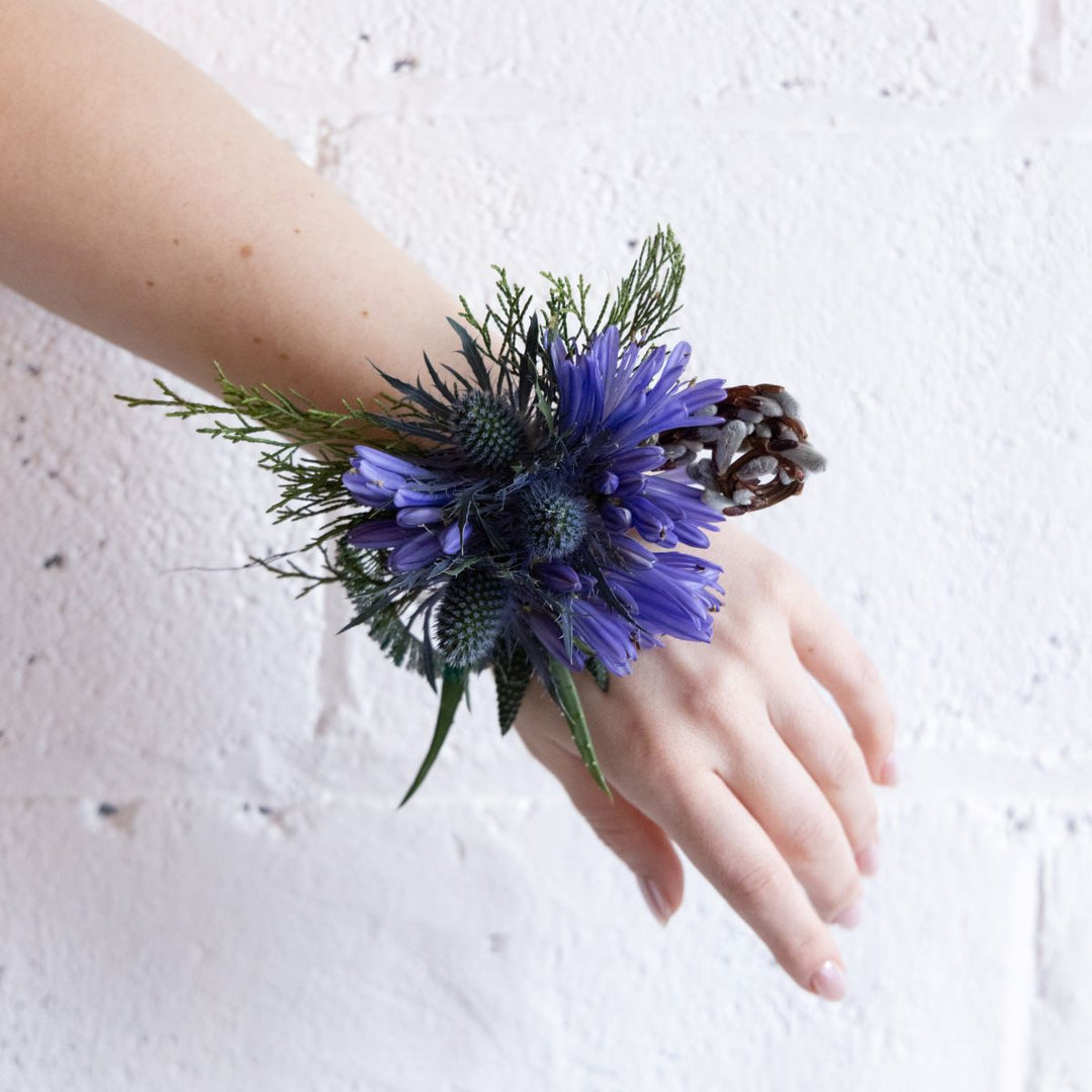 Hand wearing a wrist corsage made of purple flowers and greenery against a light background