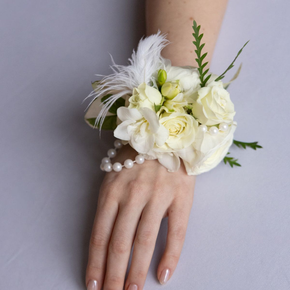 White floral corsage with feathers and pearls on a wrist against a gray background