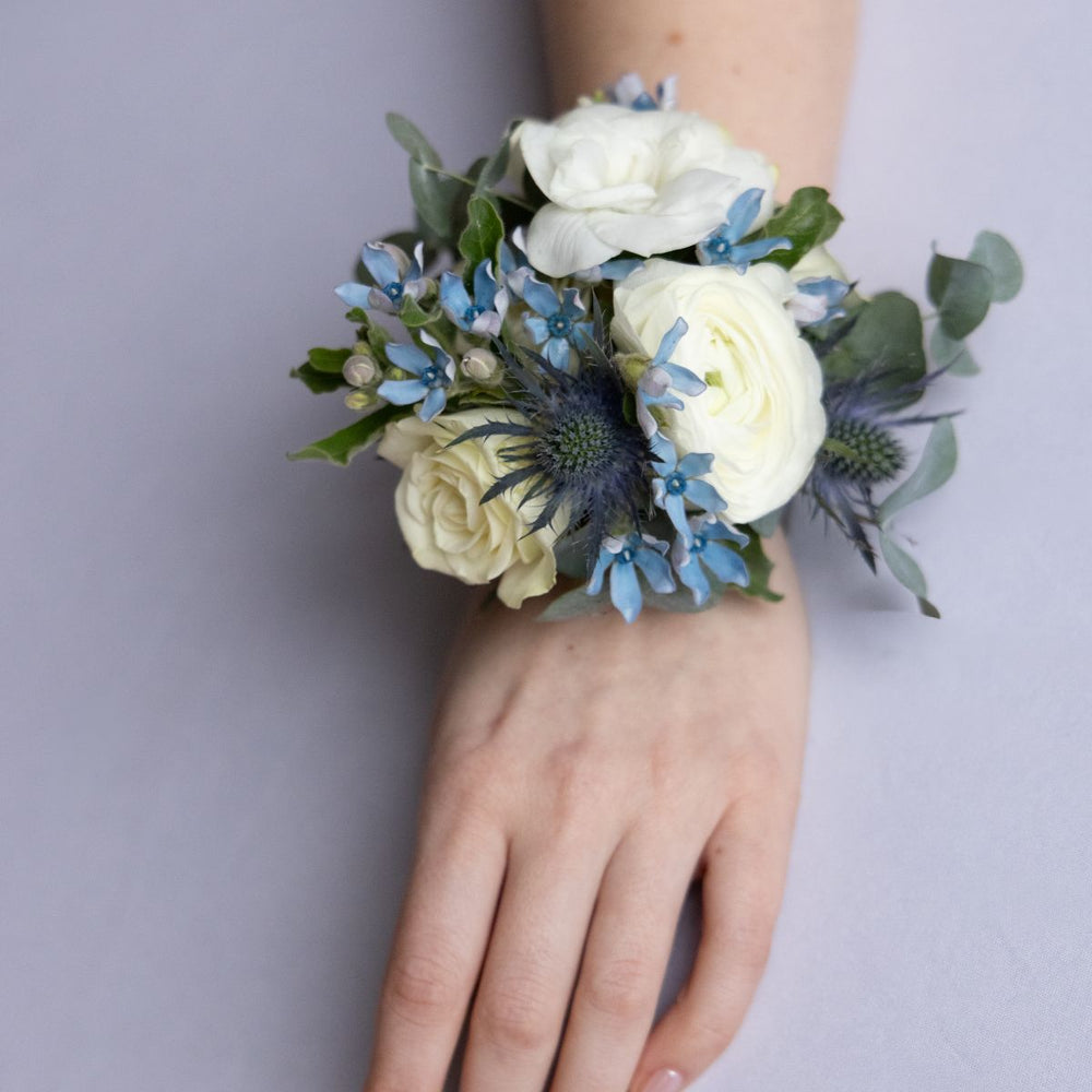 Hand wearing a wrist corsage with white and blue flowers on a plain background