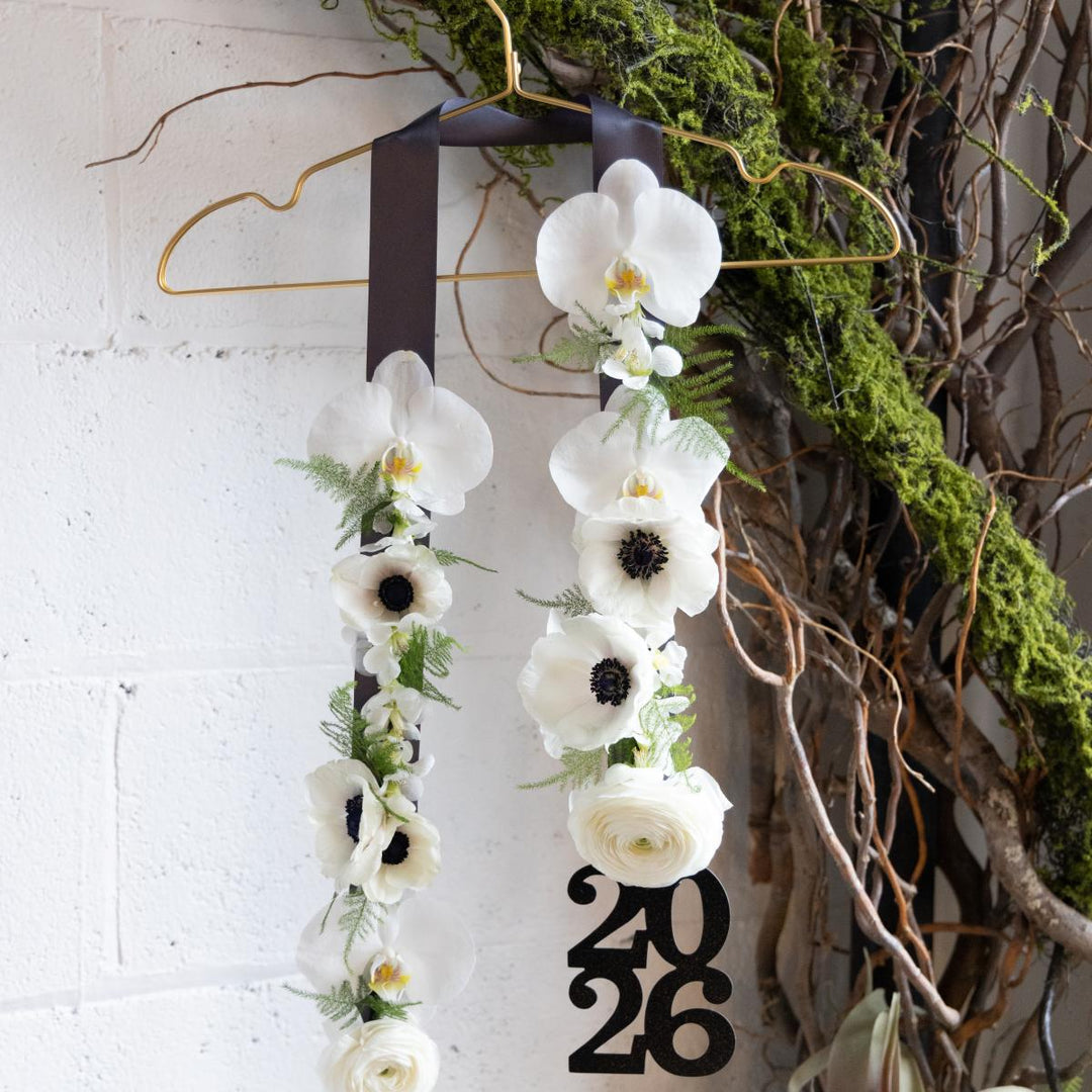 White floral graduation stole with '2026' on a hanger against a white brick wall.