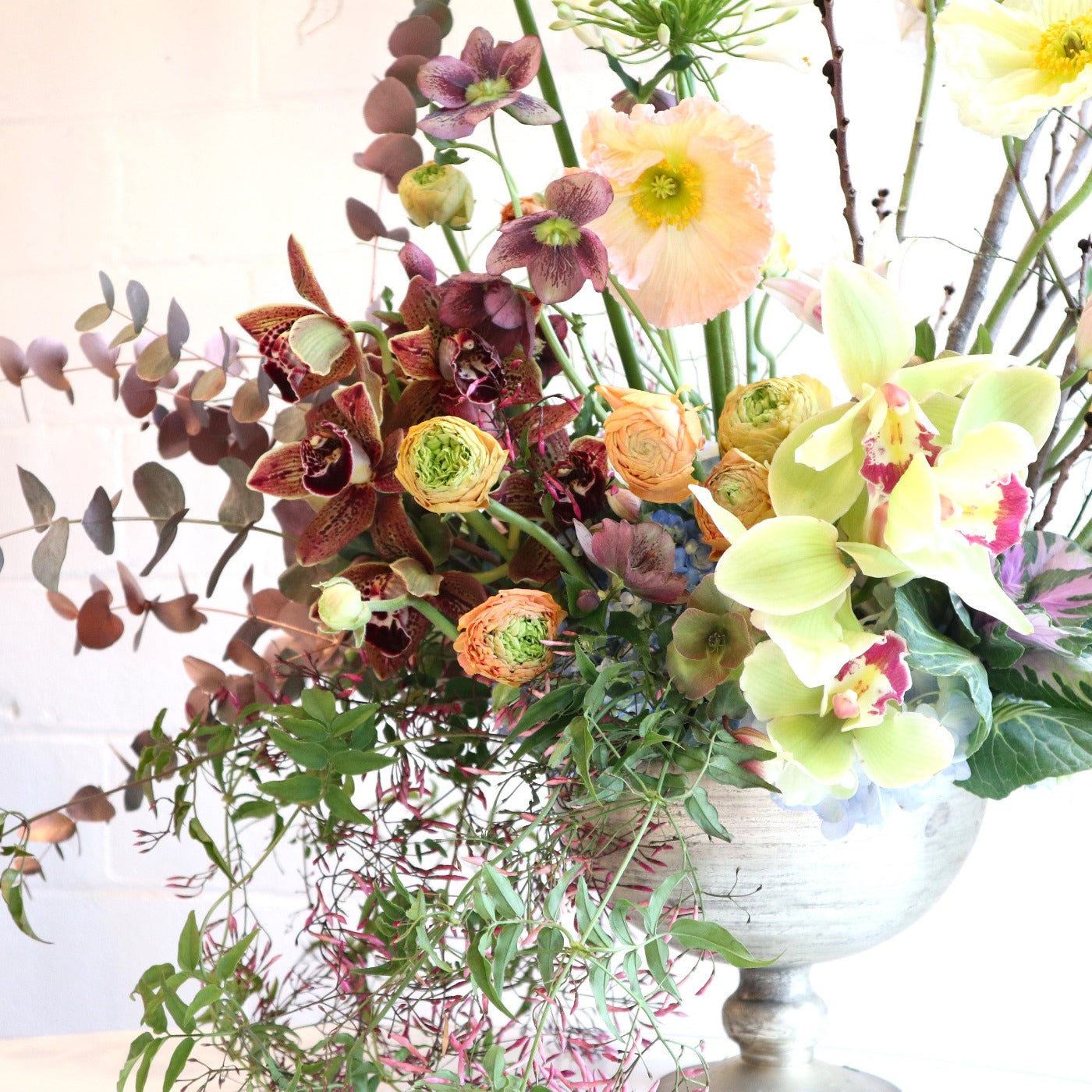 Springtime Majesty | Close up on the eucalyptus, orchis, greenery, and other florals. Colors are green, pink, purple, orange, yellow, and pink. In a reflective vase. Photo taken against a white background.