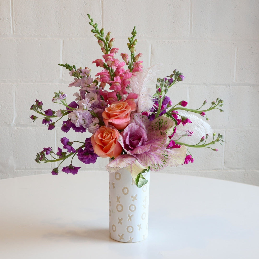 XOXO Vase | A lush arrangement with pastel purple, pink, and peach colors. Snapdragons, roses, decorative kale, feathers, and other accent florals. Arrangement is in a white vase with decorative x's and o's.