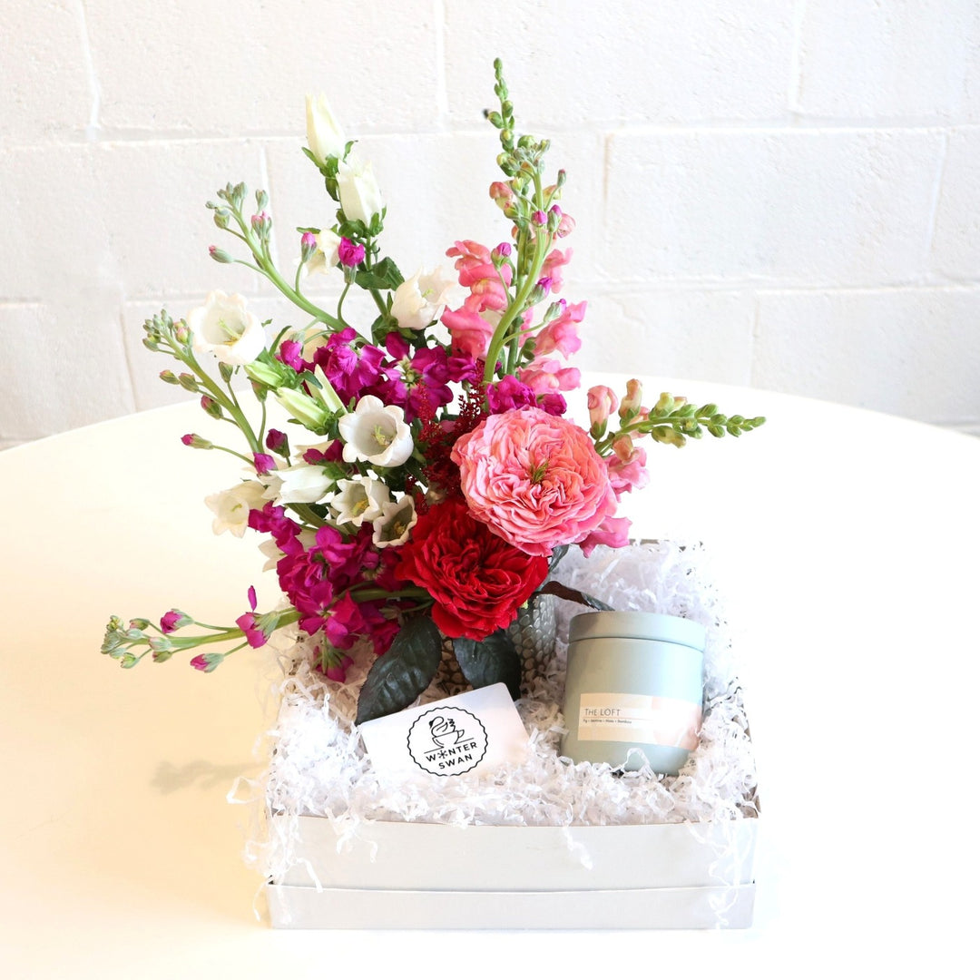 This gift box is filled with flowers, a candle and winter swan giftcard. The flower may include pink snapdragons, white campanula, pink snapdragons, garden roses and stock.