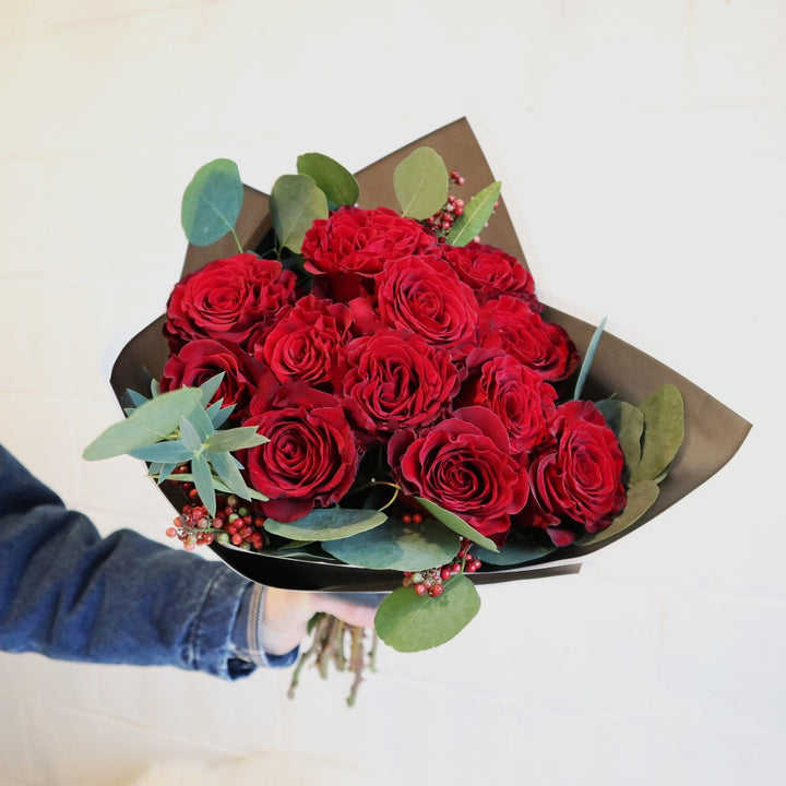 Dozen Roses Wrapped | This image includes a dozen red roses with eucalyptus greenery accentuating the outside of the roses, with everything wrapped in a black cellophane wrapping.