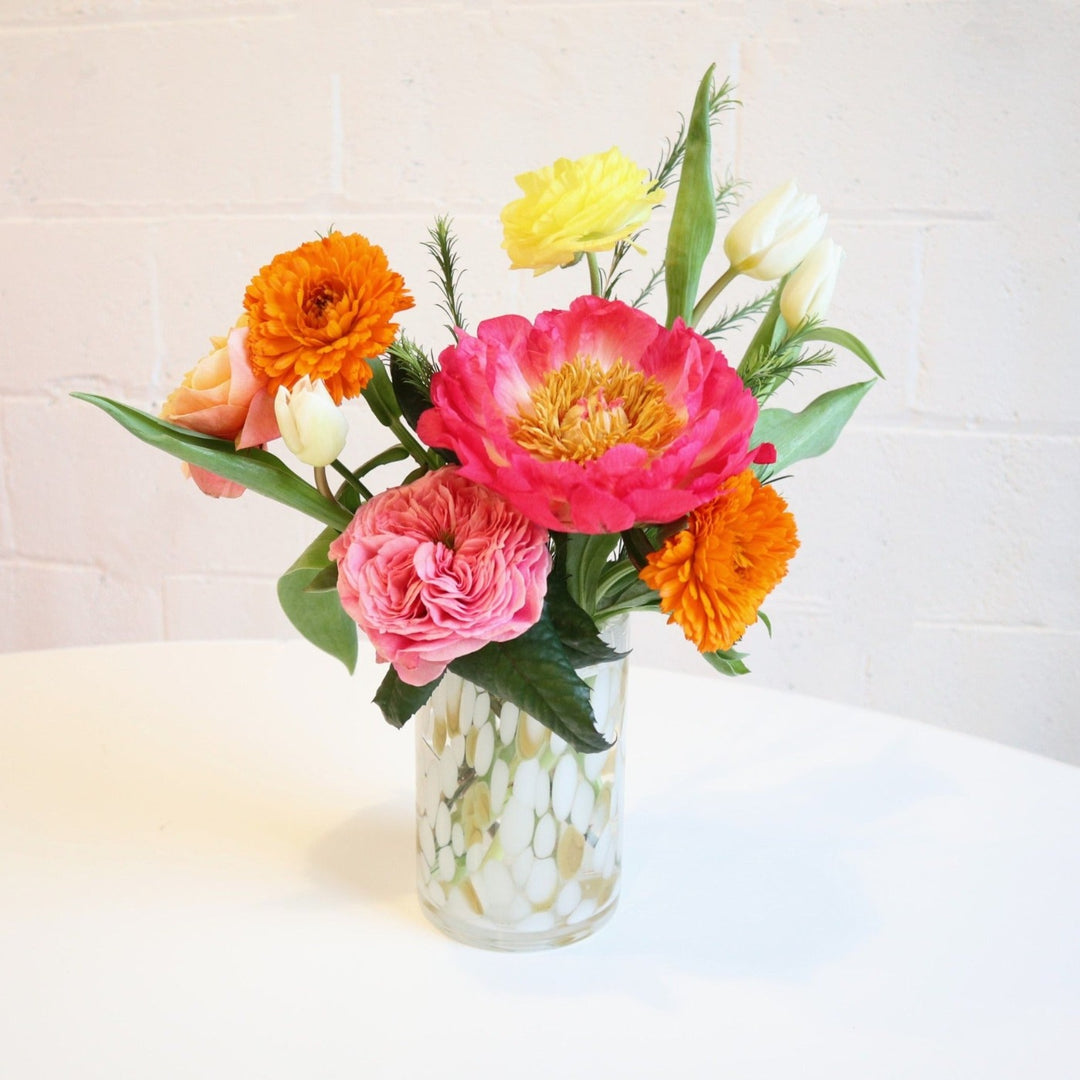 Cherish | A vase arrangement filled with one pink garden rose, one coral peony, two calendula, one orange garden rose, twi white tulips and one yellow ranunculus, in a cream and tan glass vase, on a white background.