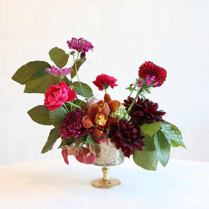 Arrangement in a gold vase with fuchsia garden roses, orchids, fall foliage, mums, and dahlias, all in a palette of deep, sophisticated colors