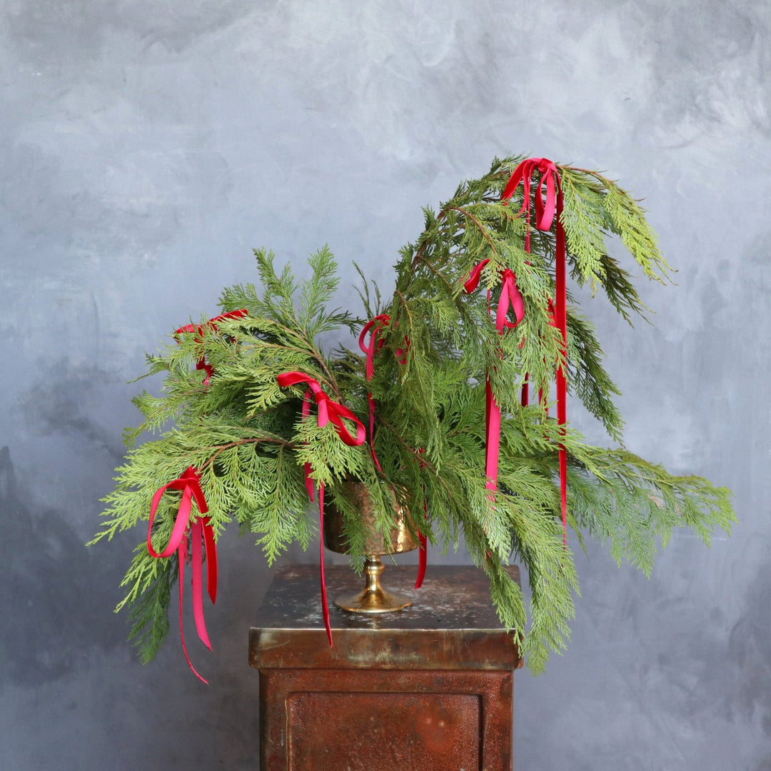 Lush evergreen branches, gracefully arranged in a vintage style brass vase. The cascading greenery is adorned with vibrant red ribbons.