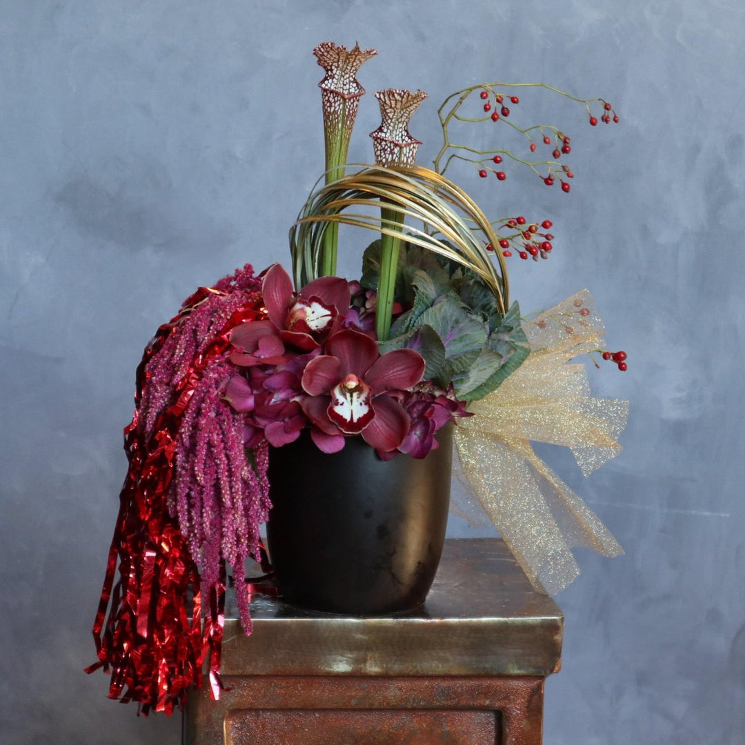 A modern floral arrangement in a black vase featuring deep red orchids, burgundy hydrangeas, tall pitcher plants, golden loops, red berry sprigs, and cascading red tinsel, accented with a sheer gold bow.