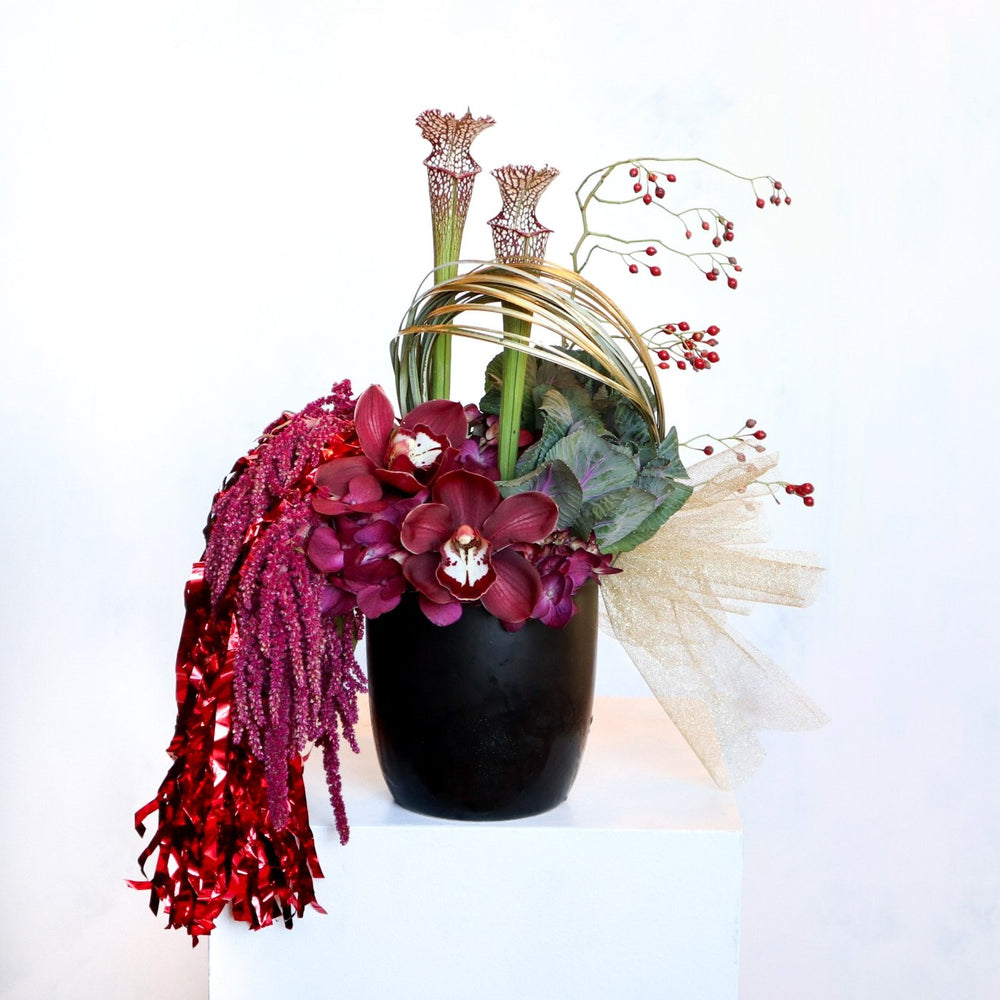 A modern floral arrangement in a black vase featuring deep red orchids, burgundy hydrangeas, tall pitcher plants, golden loops, red berry sprigs, and cascading red tinsel, accented with a sheer gold bow.