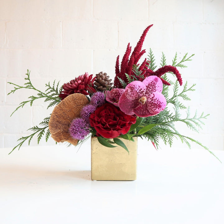 Frost | Vased Arrangement | A square gold vase with red, purple, neutral, and greens. Some flowers pictured, orchids, roses, pinecones, and allium.