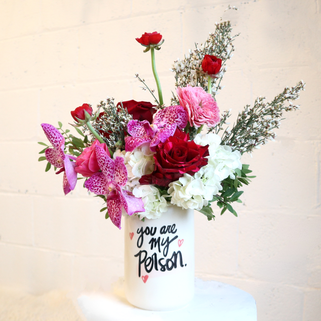 vase reads "You are my person" filled with fushia orchids, red roses, ranunculus, ginestra, white hydrangea and greenery foliage.