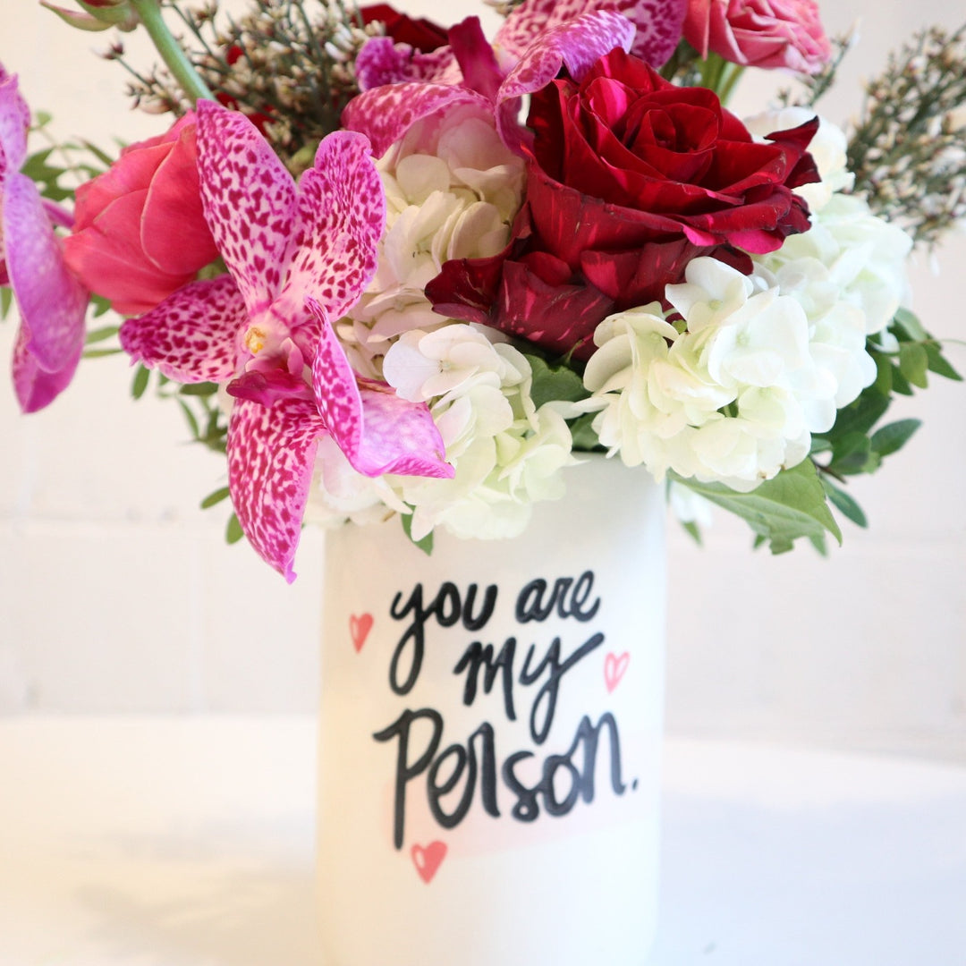 close up photo of vase, vase reads "You are my person" filled with fushia orchids, red roses, ranunculus, ginestra, white hydrangea and greenery foliage.