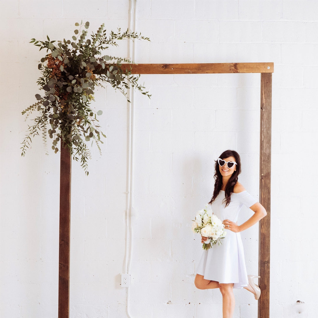 Greenery Arch Piece | A greenery arch piece with eucalyptus and other greens. Attached to a rectangle natural wood arch. In the photo a model is posing in a white dress with white sunglasses and a bridal bouquet.