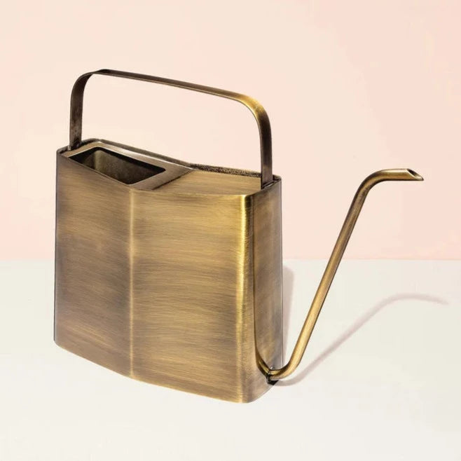 Watering Can | Modern Sprout | A Brass colored watering can with a unique tapered square shape and a long spout. Photo taken against a pink/cream background.