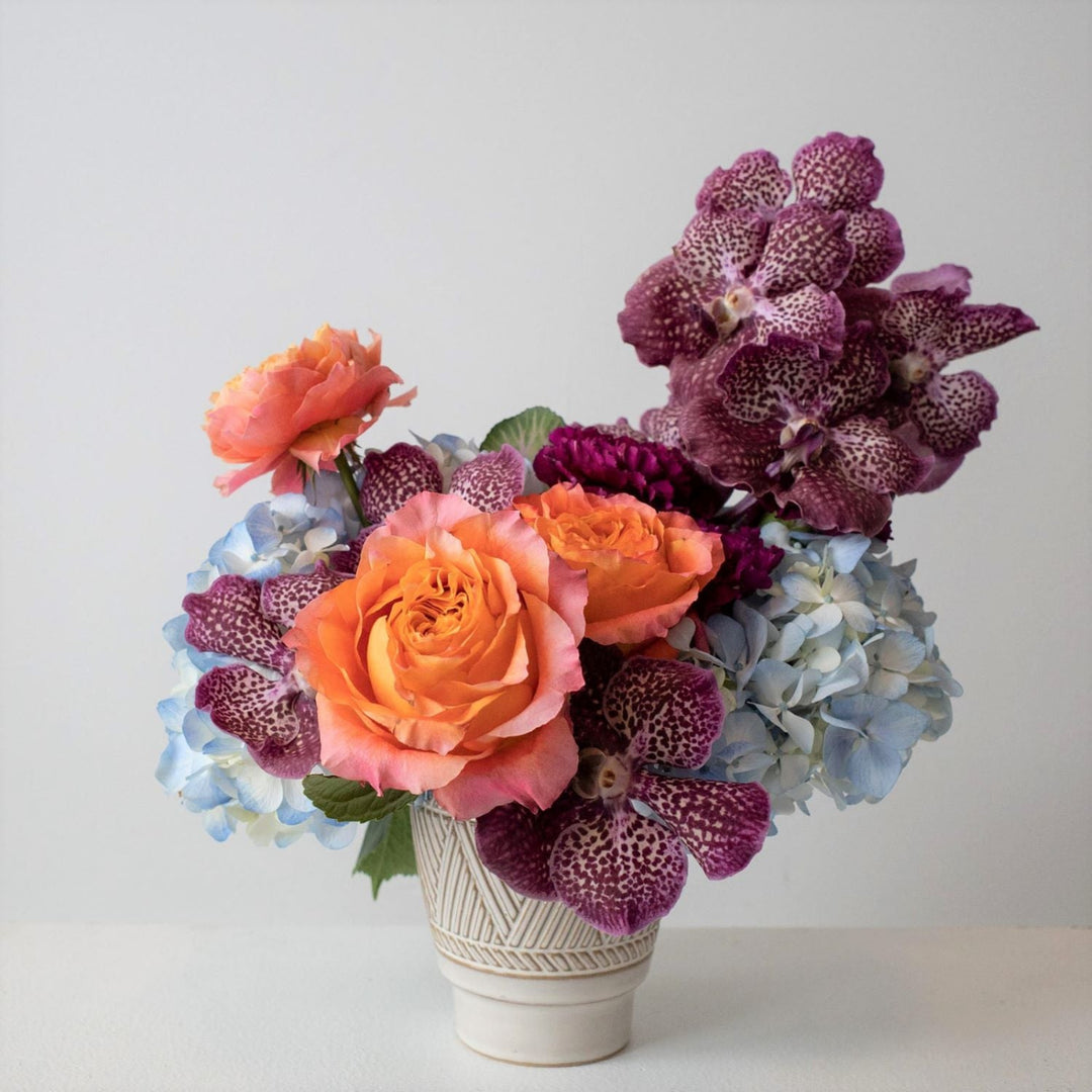 bright arrangement of hydrangea, roses, orchids  in a cream vase setting on a white table