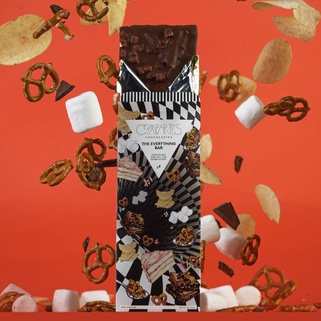A chocolate bar with Compartes branding, surrounded by various candies and marshmallows exploding around it, all against an orange background.
