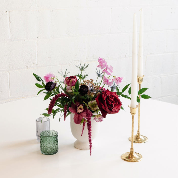 A floral centerpiece arrangement featuring a white vase, gold-colored candle holders, and a selection of purple, pink, and white flowers, placed on a white surface with a plain background.