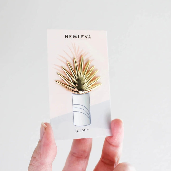 Hemleva Enamel Pins | Gold fan palm pin with a shiny reflective surface. Package is bein held against a white backdrop.