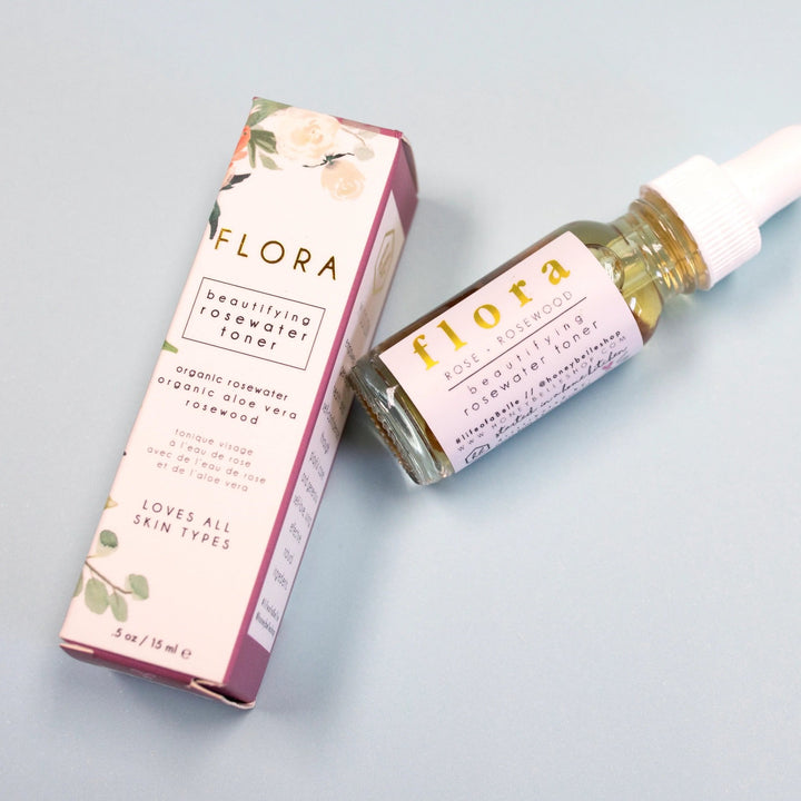 Flora: Rosewater Toner | Honey Belle | Beautifying rosewater toner, organic rosewater, organic aloe vera, rosewood, loves all skin types. .oz/15 ml. White packaging with a floral pattern and purple accents.