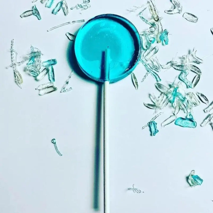 Good Lolli Blue Razz Lollipop. Transparent blue lollipop against light backdrop with blue/clear sugar shards placed decoratively around.