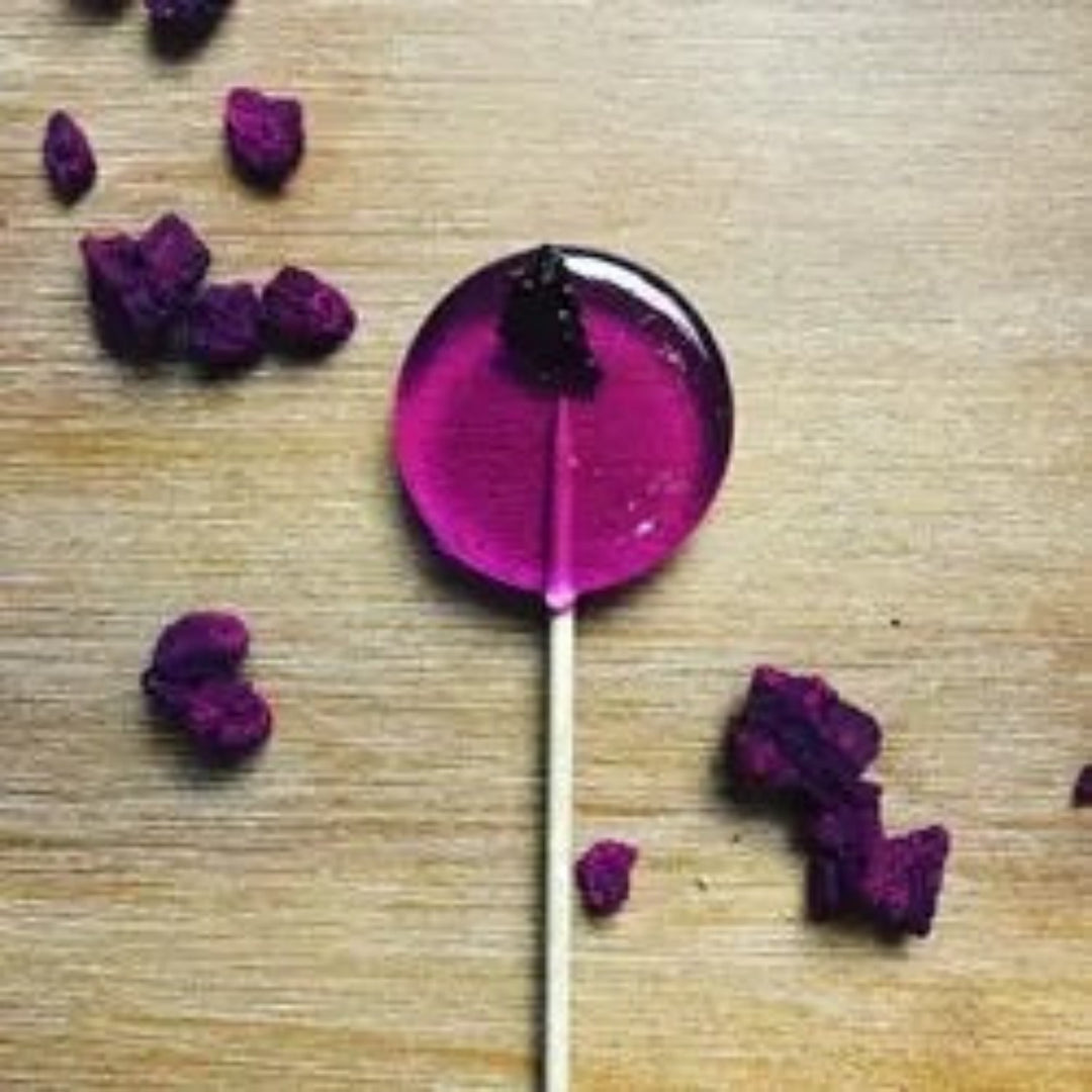 Good Lolli Dragonfruit Lollipop. Purple/Magenta color lollipop with dragonfuit pieces, photo taken against wooden backdrop with dragonfuit pieces placed decoratively around.