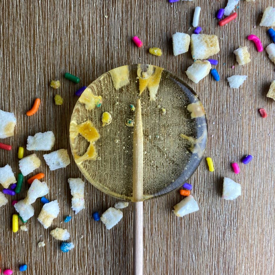 A birthday cake lollipop with candy cane pieces and colorful sprinkles on a wooden surface.