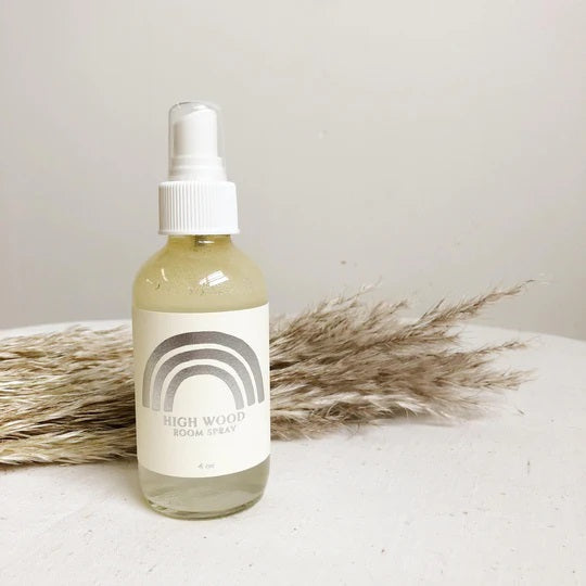High Wood Room Spray | A simple white label with a silver rainbow on a glass spray bottle. Decorative grasses placed in the background.