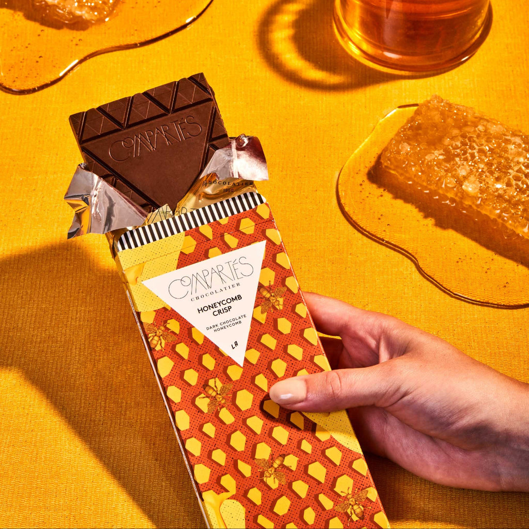 A gourmet dark chocolate bar with honeycomb pieces, presented on a yellow background with a glass of milk and more honeycomb to the side.