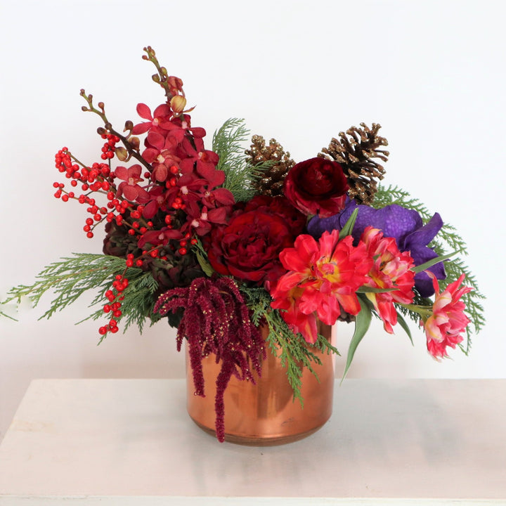 Arrangement in a rose gold vessel, with pink tulips, red roses, red mokara roses, burgundy ranunculus, vanda orchids, red amarathus, pinecones, red ilex berries, wintergreens. Taken on a gray background.