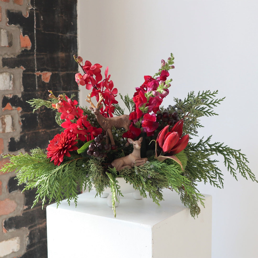Arrangement in a white vase, with small round feet. Filled with red oncidium orchids, burgundy snapdragons, red amaryllis, red mum and cedar evergreen.