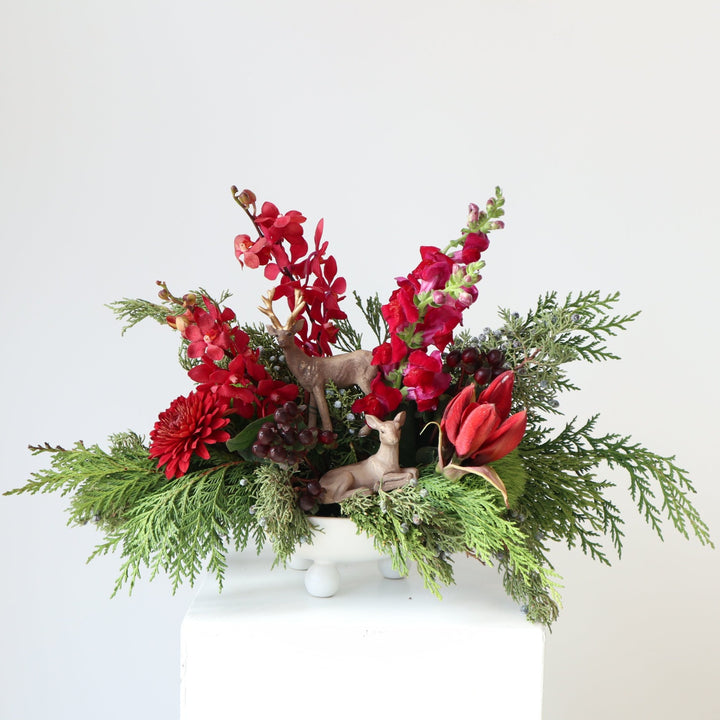 Arrangement in a white vase, with small round feet. Filled with red oncidium orchids, burgundy snapdragons, red amaryllis, red mum and cedar evergreen. 