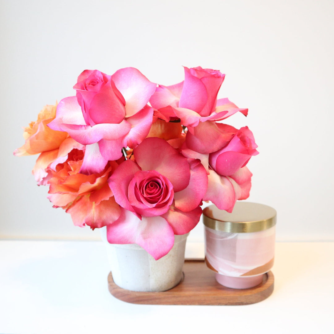 An arrangement with pink roses and other seasonal blooms in a white striped vase on a wooden base with a pink candle that has a gold lid.