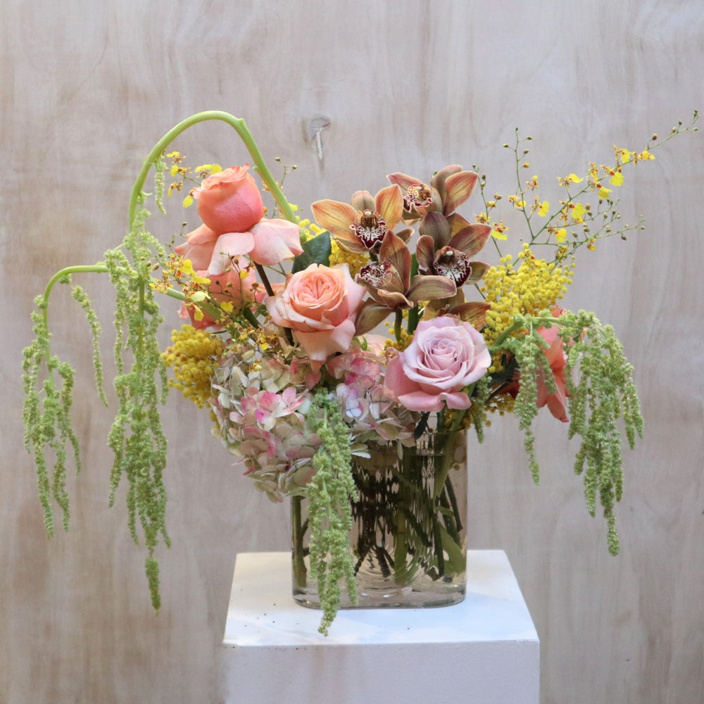 Sunbeam | An arrangement featuring roses, orchids, hydrangea, ameranthus, and other florals. Colors or gree, pink, and yellow. In a transparent vase. Photo taken against wooden background.