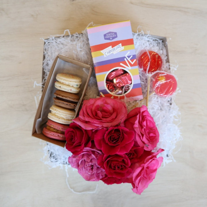 A light colored background with two cream colored macarons and two lolli pops and a seattle chcocolate truffle box. in a white gift box with a pink rose bouquet at the bottom.