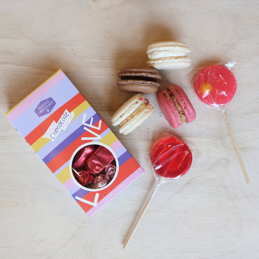 A light colored background with two cream colored macarons and two lolli pops and a seattle chcocolate truffle box.