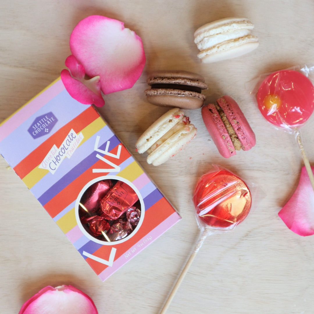 A light colored background with two cream colored and one pink macarons and two lolli pops and a seattle chcocolate truffle box.