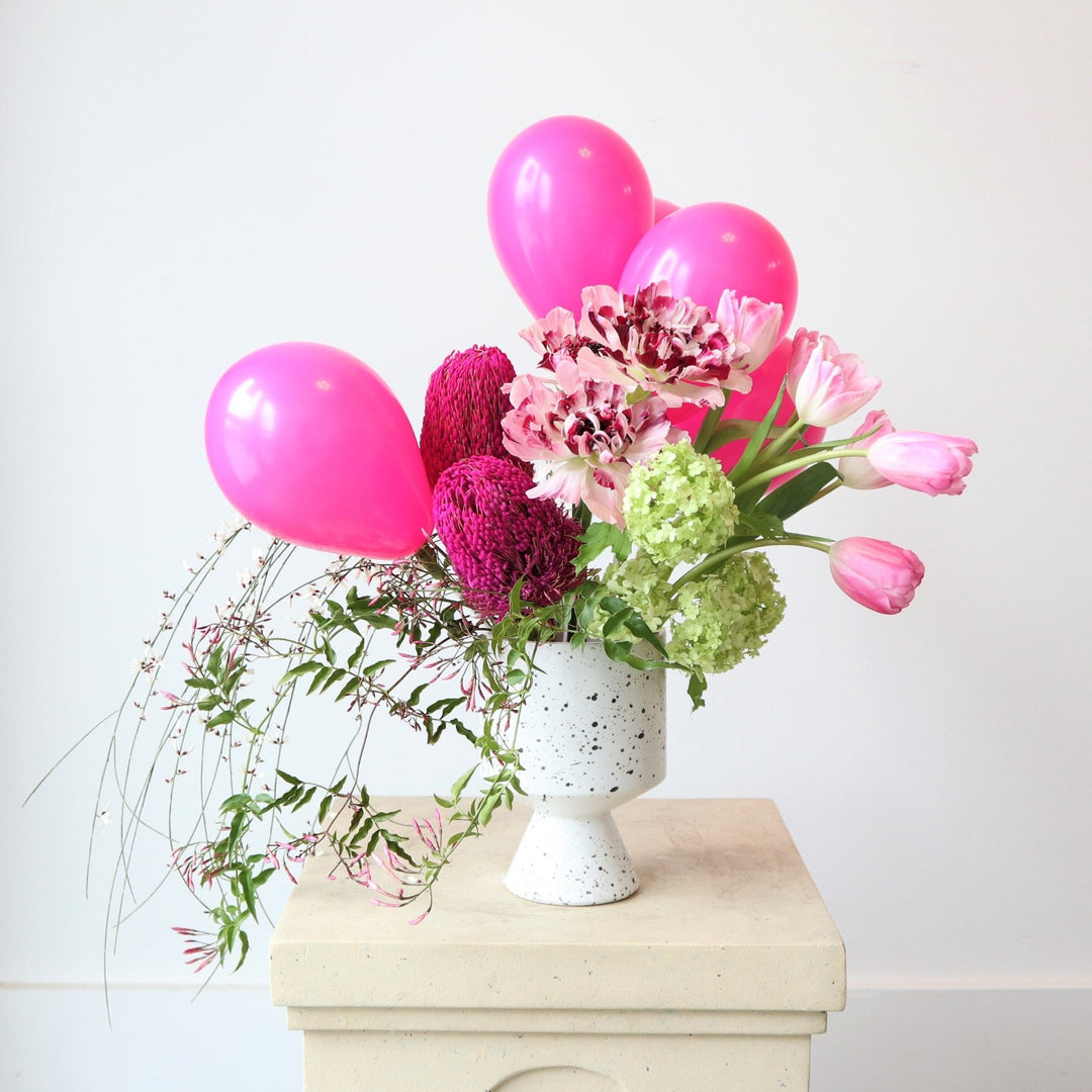 Cheerful | A bright pink and green arrangement featuring pink balloons, tulips, green hydrangea, greens, and other florals. Vase is white with black speckles. Photo taken on a pedestal against a white background.