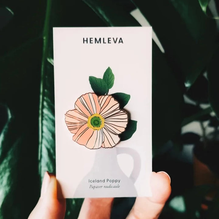 Hemleva Enamel Pins | Iceland Poppy, Papaver nudicaule. A pink poppy pin with a yellow and green center. Package is being held up against a leafy background.