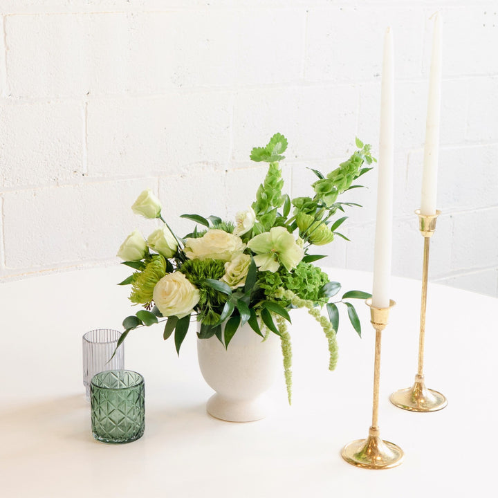 A floral centerpiece arrangement featuring a white vase, gold-colored candle holders, and a selection of purple, pink, and white flowers, placed on a white surface with a plain background.