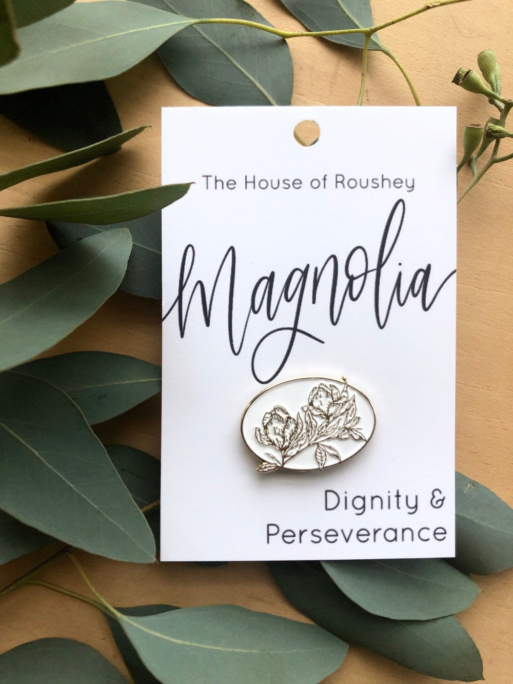 Enamel Pins by Christy Roushey | The House of Roushey, Magnolia pin. It is a white pin in an oval shape with magnolia flowers outlined with fine lines. The bottom of the label reads "Dignity & Perseverance".
