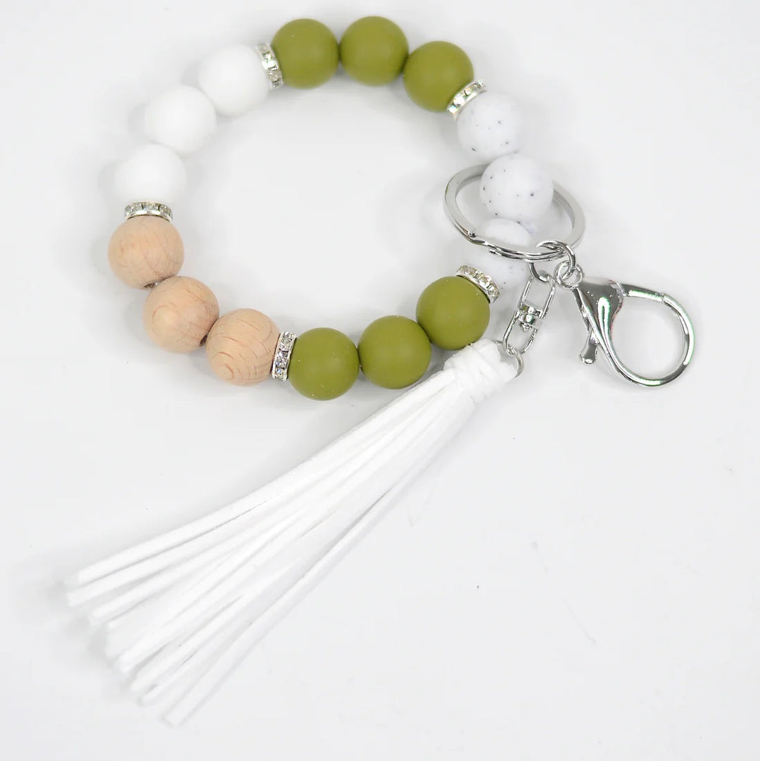 Assorted Beaded Keychains | A natural and olive colored keychain bracelet. There are white, olive green, marbled, and wooden beads. Accented with a silver key ring and white tassel.