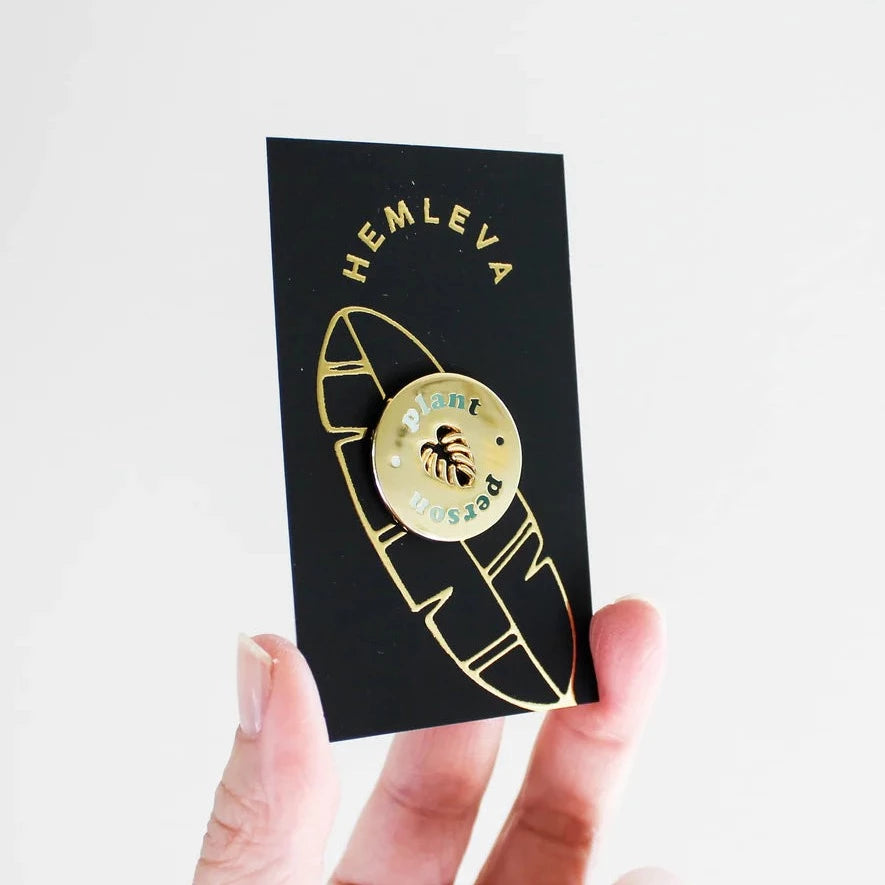 Hemleva Enamel Pins | Gold "Plant Person" pin with a cutout of a monstera leaf in the middle. Black and gold packaging.