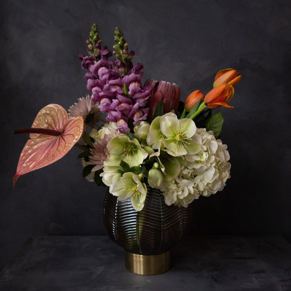 Dark gray background with vased arrangement that has green helleborus, lilac colored mums, purple snapdragons, orange tulips and a peach colored anthurium.