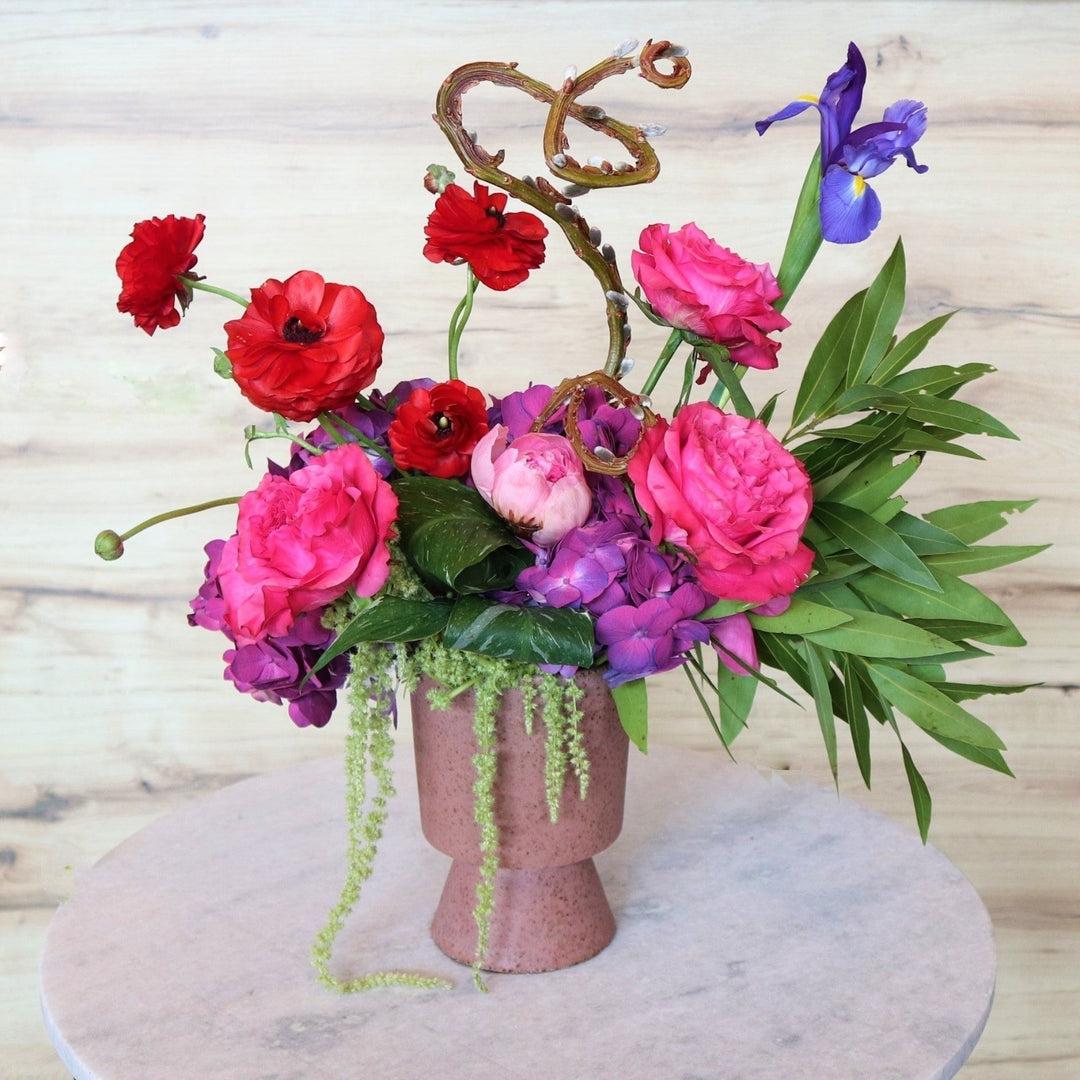 Light background with Floral arrangement in front. Deep pink vase filled with pink roses, red ranunculus, one iris, purple hydrangea, green amaranthus, greenery, pussy willow and one pink peony.